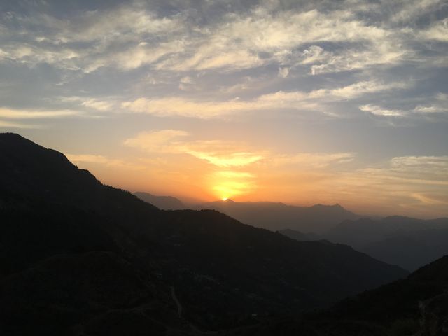 This image captures a tranquil mountain sunset with the sun disappearing behind scenic hills. The sky is adorned with a gentle blend of orange and blue hues, creating a tranquil scene perfect for projects highlighting nature's beauty, serenity, and travel destinations. Ideal for use in travel blogs, magazines, and promotional materials for eco-tourism.