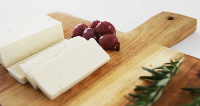 Slices of fresh feta cheese are arranged on a wooden chopping board next to a small bunch of red grapes. A sprig of rosemary is placed nearby, adding a touch of green. This scene is perfect for use in culinary blogs, gourmet food websites, marketing materials for cheese products, or publications focusing on healthy eating and snack ideas.