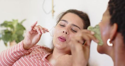 Female friends enjoying skincare ritual using facial roller and gua sha on cozy sofa