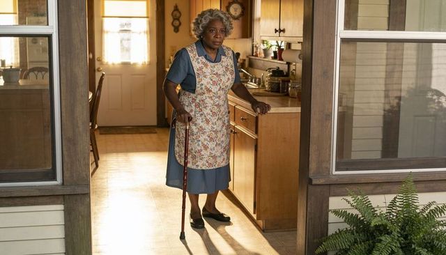 Senior woman steadying in sunlit kitchen doorway holding wooden cane wearing floral apron