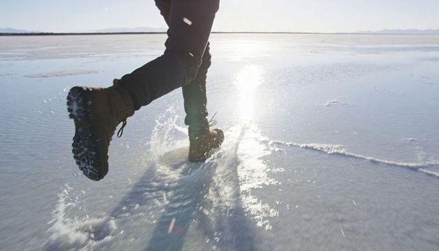Boots striding across reflective salt flat splashing water with sun glare and motion