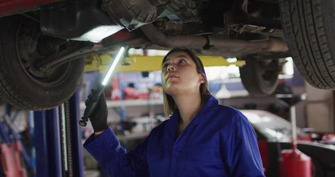 Female mechanic inspecting car underside with led inspection light on lift in workshop
