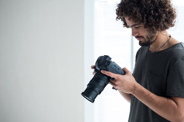 Male photographer with curly hair reviewing captured photos on his digital camera in a studio. Ideal for use in articles about photography, creative professions, camera equipment reviews, or modern technology in creative industries.