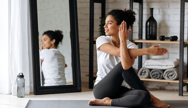 Young woman practicing seated spinal twist on yoga mat by mirror in bright home studio