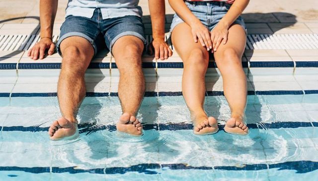 Young couple sitting on pool edge dipping feet in clear water under bright summer sun