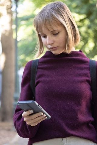 Young woman checking phone on park path wearing maroon sweater and backpack