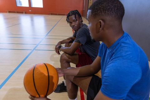 African american teammates sitting on gym bench holding basketball discussing strategy