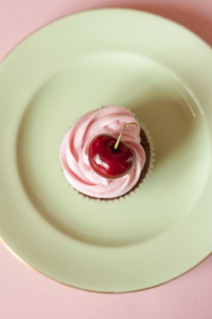 Cherry cupcake with pink frosting and a cherry on top, placed on light green plate. Ideal for use in food blogs, recipe books, bakery advertisements, or dessert menus. Represents sweet indulgence, attractiveness, and culinary presentation.