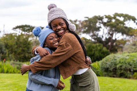 African american sisters hugging and smiling in park wearing cozy knit beanies
