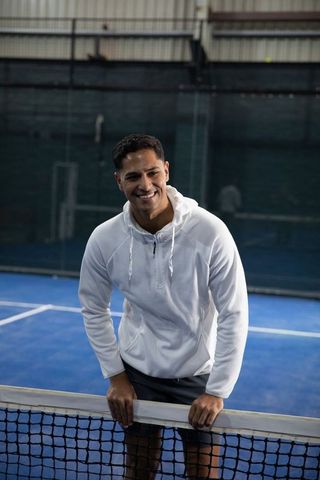 Smiling man leaning on tennis net on indoor blue court wearing white hoodie and dark shorts