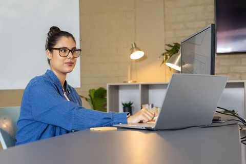 Asian woman working at minimalist desk typing on laptop with dual monitor and warm desk lamp