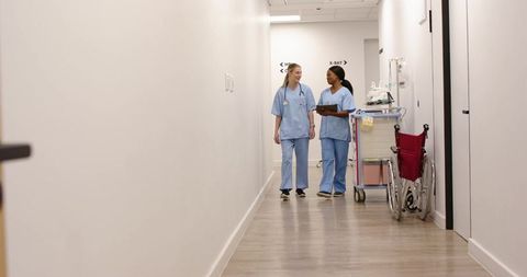 Clinic nurses walking and consulting in corridor with mobile medical cart and wheelchair