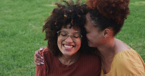 African american female couple embracing and smiling on park grass, candid outdoor love