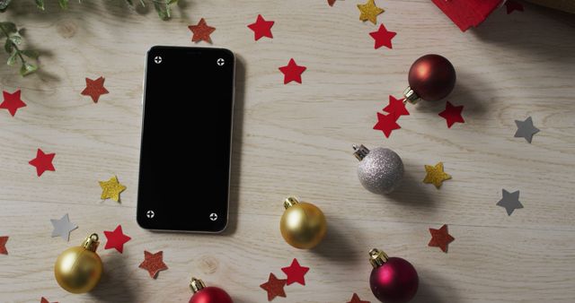 Smartphone placed on wooden table amid colorful Christmas ornaments and star-shaped confetti. Suitable for holiday-themed promotions, technology integration in celebrations, and festive marketing campaigns.