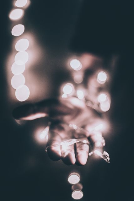 Hand holding fairy lights with a dark background creating a magical and dreamy ambiance. Bokeh effect of lights blurring in background enhances festive feel. Suitable for use in holiday, celebration, and party themes, as well as artistic and inspirational content.