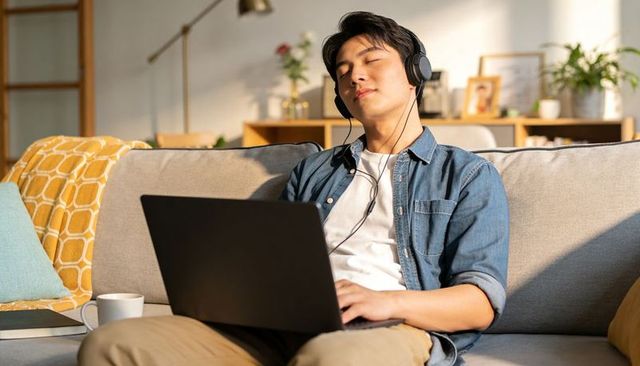 Asian man relaxing with headphones and laptop on cozy living room sofa for remote work