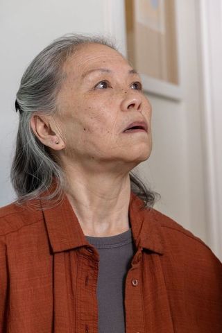 Senior asian woman gazing upward in contemplative serene indoor portrait wearing rust shirt