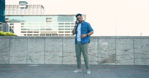 Man standing on urban terrace talking on smartphone adjusting denim shirt