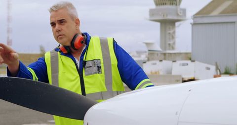 Ground crew member closely inspects an aircraft propeller at an airport wearing high-visibility vest and headset. Focuses on ensuring the safety and readiness of the plane. Ideal for use in aviation industry materials, safety procedures guides, and technical training content highlighting maintenance practices.
