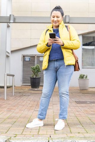 Mature woman standing on urban plaza checking smartphone in yellow puffer jacket and jeans