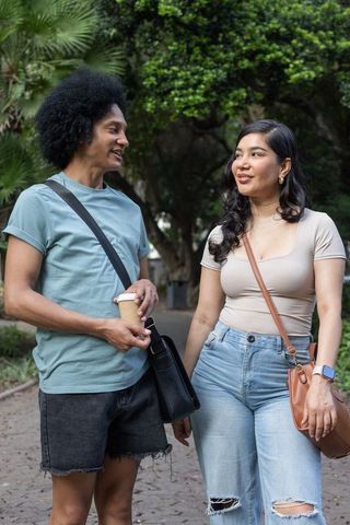 Multiracial couple walking and chatting on tree-lined urban park path with coffee