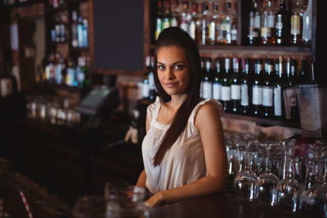 Young female bartender standing confidently at a bar counter with bottles and glasses in the background. Ideal for use in articles or advertisements related to nightlife, hospitality, bar services, and professional bartending.