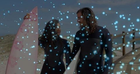 Couple carrying surfboards on beach boardwalk wearing wetsuits with blue bokeh overlay