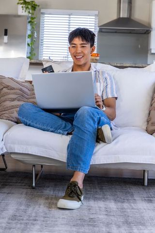 Asian man smiling while online shopping with laptop and credit card on cozy modern sofa