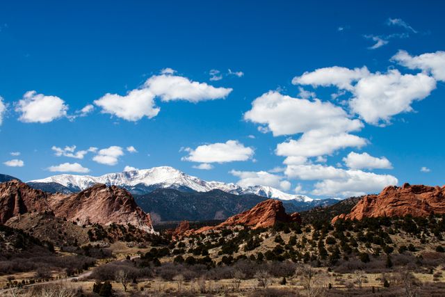 Rugged red rock formations stand in contrast against snow capped mountains under a bright blue sky with white clouds. This breathtaking natural landscape captures the stark beauty of arid climate regions and is ideal for illustrating themes related to outdoor adventures, geology, travel destinations, and nature exploration.