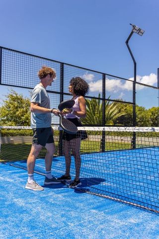 Mixed-race couple chatting at net on bright blue padel court holding paddles