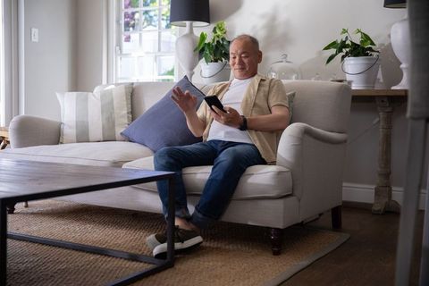 Mature asian man using smartphone and gesturing on beige sofa in bright modern living room