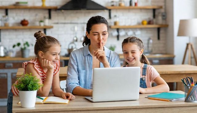 Mother shushing daughters while studying at laptop in cozy kitchen workspace