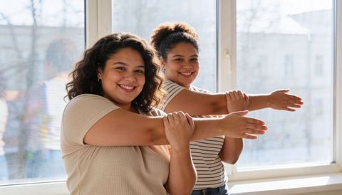 Smiling women stretching arms across chest in sunny window light, casual fitness