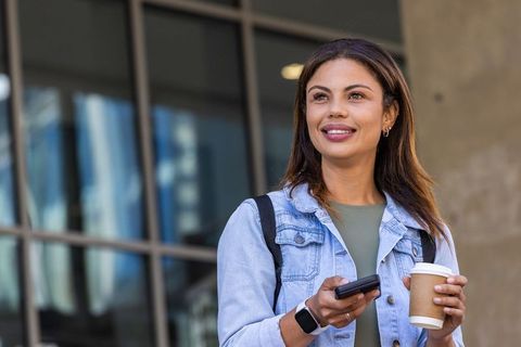 African american woman holding coffee and smartphone, smiling outside glass building