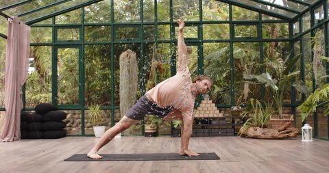 Practicing side angle yoga pose in glass greenhouse surrounded by tropical plants