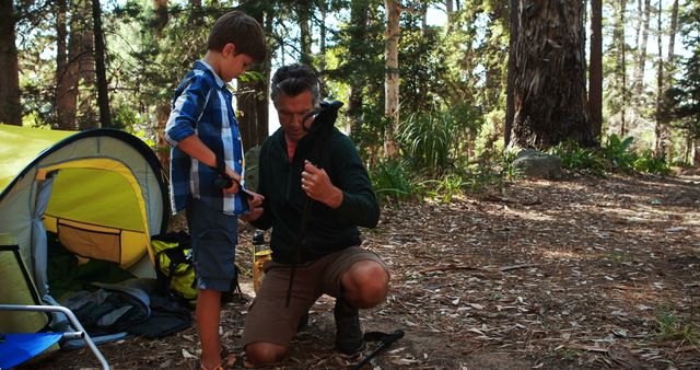 Father and son spending quality time together on a camping trip, working together to set up their tent in a wooded forest. Perfect for depicting family bonding, outdoor activities, teamwork, and love for nature.