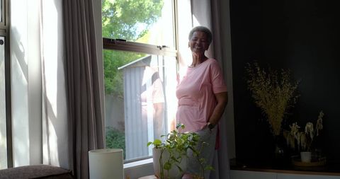 Smiling senior african woman standing by sunlit window caring for houseplant, relaxed at home