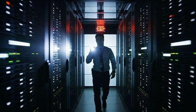 Backlit technician walking through server aisle holding flashlight in secure datacenter