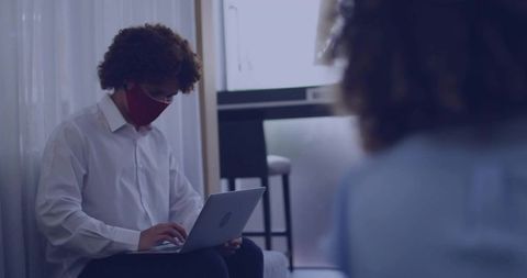 Young professional working remotely on laptop wearing face mask in modern living room