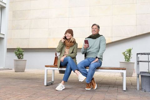Middle-aged couple relaxing on urban bench using smartphones and planning weekend outing