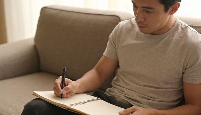 Man writing in spiral notebook on sofa focusing on study, journaling and planning