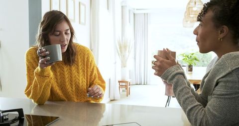 Two women chatting and sipping coffee at bright kitchen island in cozy knit sweaters
