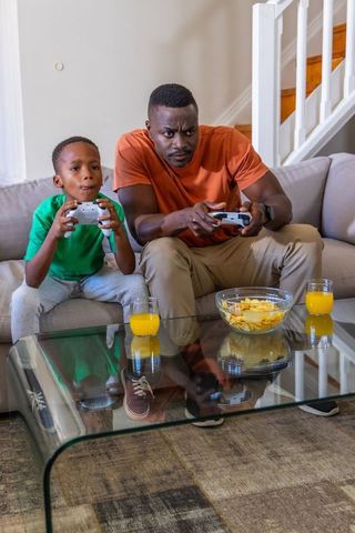 African American father and son playing console game on sofa, sharing snacks and juice