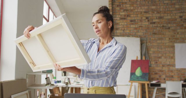 Female artist in casual striped shirt inspecting a blank canvas with focus. Bricked-walled art studio with paintings and easels visible in background. Ideal for illustrating concepts of art creation, artistic process, creativity, and professional artists at work.