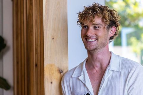 Smiling curly-haired man leaning on light wood column wearing white shirt and sunglasses