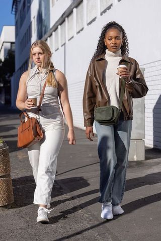 Sunlit urban friends walking with coffee wearing brown leather jacket and crossbody bags