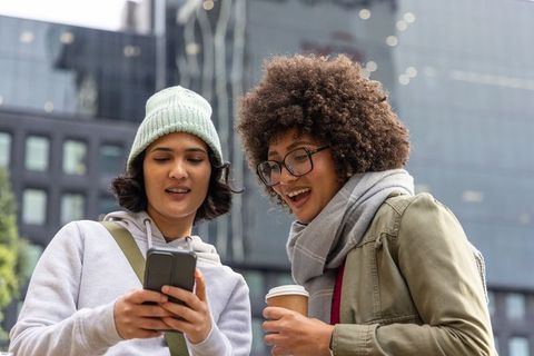 Multicultural friends checking smartphone in urban plaza holding coffee, casual street style