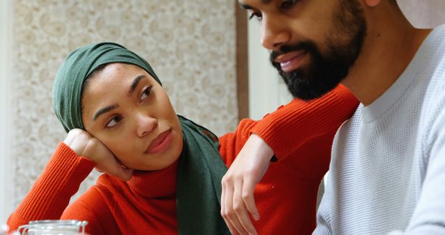 Couple sitting indoors having a thoughtful conversation, possibly addressing important relationship issues. Woman wears green headscarf and orange sweater while a bearded man listens attentively. Ideal for use in articles about relationships, communication in marriage, or emotional support.