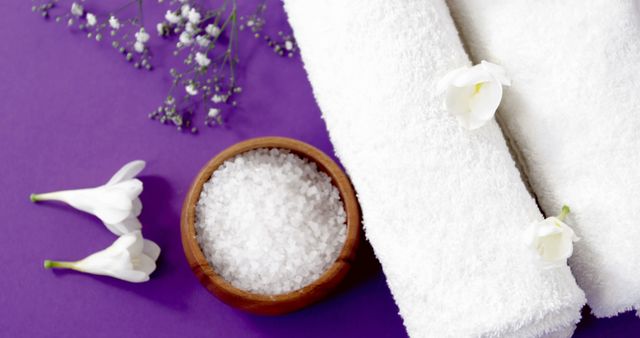 A serene spa setup features fluffy white towels, a wooden bowl of salt crystals, and delicate white flowers on a vibrant purple background, with copy space. The composition evokes a sense of relaxation and is ideal for wellness and beauty themes.