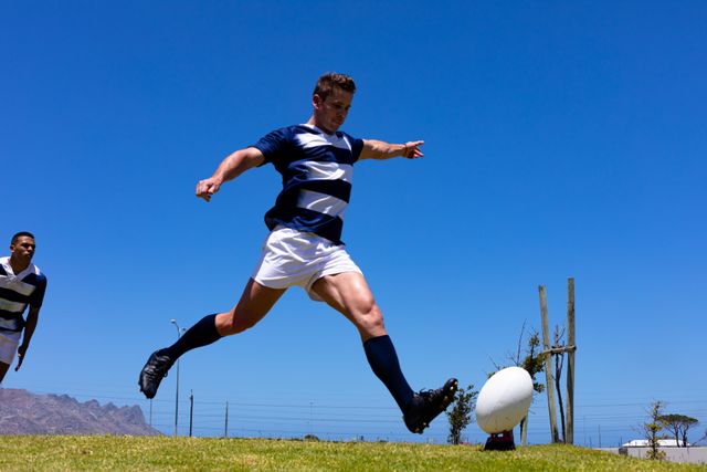 Caucasian male rugby player kicking ball during a game on a sunny day, with teammate in background. Ideal for sports-related content, athletic competition promotions, teamwork and dynamic action visuals, and outdoor activity advertisements.